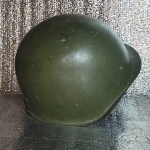 Puede incluir: Un casco militar verde con un acabado ligeramente descolorido. El casco tiene una forma redondeada y una superficie lisa.