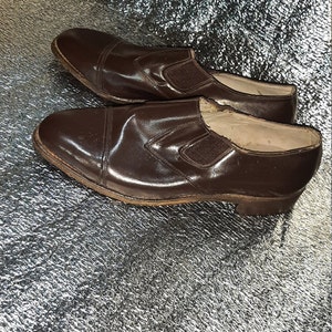 May include: A pair of brown leather slip-on shoes. The shoes have a rounded toe and a small elastic panel on the side. The leather has a glossy finish. The soles are a lighter brown color.