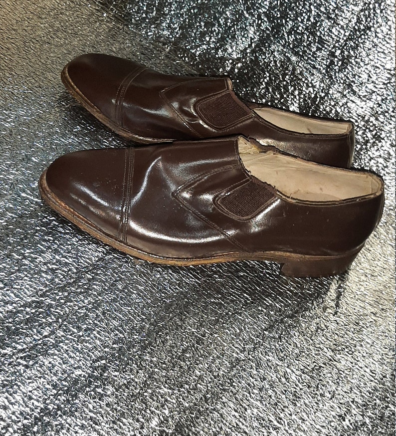May include: A pair of brown leather slip-on shoes. The shoes have a rounded toe and a small elastic panel on the side. The leather has a glossy finish. The soles are a lighter brown color.