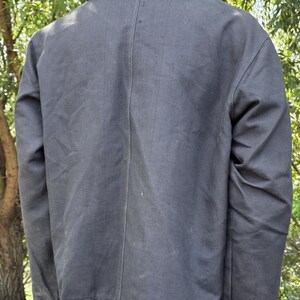 K&ouml;nnte beinhalten: Dunkelgraue Jacke mit Kragen und gekn&ouml;pften Manschetten. Die R&uuml;ckseite der Jacke ist sichtbar und zeigt eine vertikale Naht in der Mitte. Die Jacke scheint aus einem strapazierf&auml;higen Stoff gefertigt zu sein.