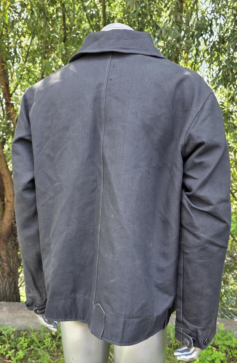 K&ouml;nnte beinhalten: Dunkelgraue Jacke mit Kragen und gekn&ouml;pften Manschetten. Die R&uuml;ckseite der Jacke ist sichtbar und zeigt eine vertikale Naht in der Mitte. Die Jacke scheint aus einem strapazierf&auml;higen Stoff gefertigt zu sein.