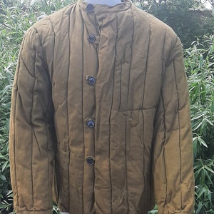 Puede incluir: Una chaqueta acolchada de color verde oliva oscuro con cuello alto y cierre de botones. La chaqueta tiene líneas de acolchado verticales y un bolsillo de parche en el lado izquierdo.