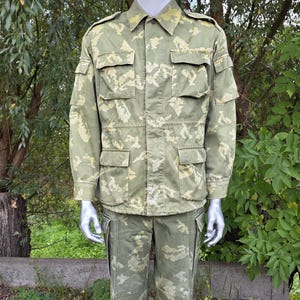 Op de afbeelding: Een jas met lange mouwen en knopen en een bijpassende broek met een digitaal camouflagepatroon in groen en beige. De jas is voorzien van een kraag, schouderkleppen en vier voorzakken met knoopsluitingen.