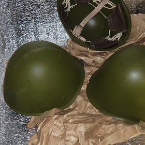 May include: Three green military helmets, two are laying flat and one is standing upright. The helmets are on a brown paper background.