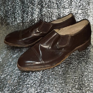 May include: A pair of brown leather slip-on shoes. The shoes have a rounded toe and elastic side panels. The soles show signs of wear. The shoes are a classic style.