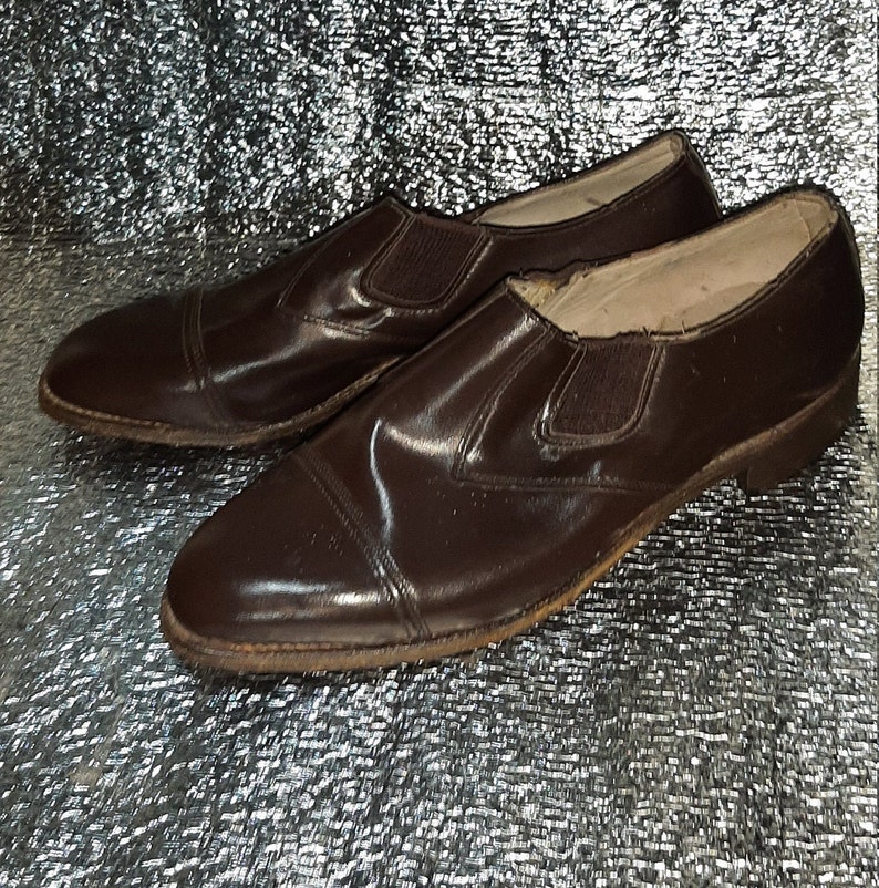 May include: A pair of brown leather slip-on shoes. The shoes have a rounded toe and elastic side panels. The soles show signs of wear. The shoes are a classic style.