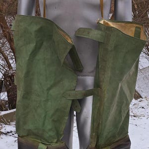 Op de afbeelding: Een paar groene waterdichte beenbeschermers met verstelbare banden en zwarte rubberen laarzen. De beenbeschermers zijn gemaakt van een dik, duurzaam materiaal en zijn ontworpen om je benen droog te houden in natte omstandigheden.