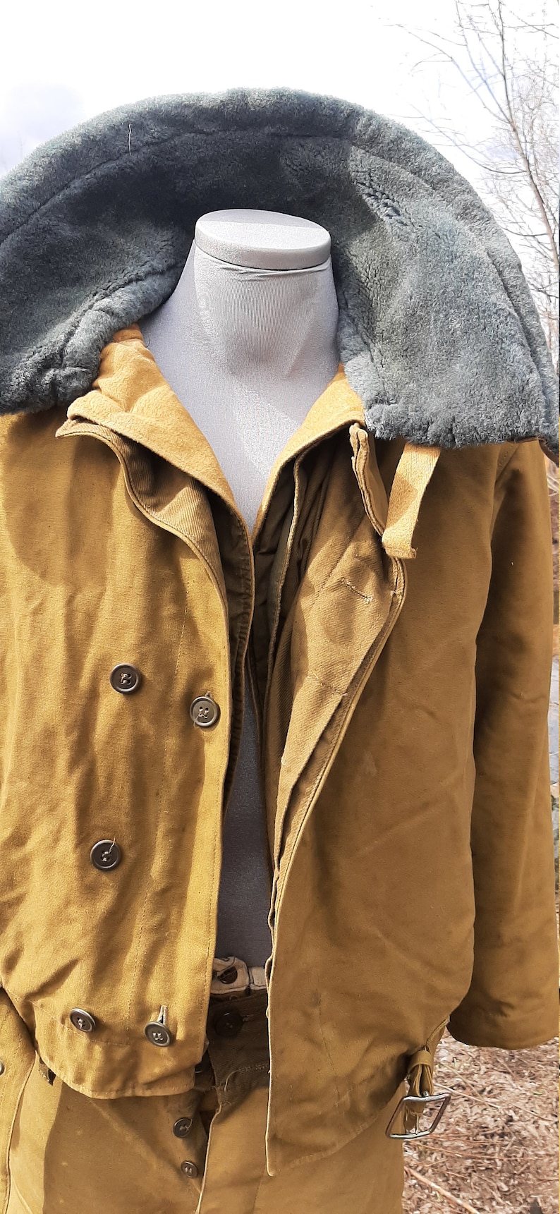 Op de afbeelding: Een bruin, gevoerd jack met een grijze bontkraag en een knoopsluiting. Het jack heeft een riem en is gemaakt van een dikke, duurzame stof.