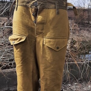 Op de afbeelding: Een broek van bruin canvas met een hoge taille en meerdere zakken. De broek heeft een knoopsluiting en een riemlus. De broek is gemaakt van een dik, duurzaam materiaal.