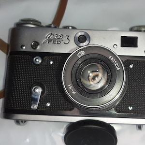 May include: A silver Fed 3 camera with a black leather strap and a black lens cap. The camera has a black lens with the text "FED" and "19-IV" on it. The lens also has the number "885206" printed on it.