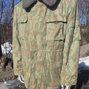 Op de afbeelding: Een camouflagejas met een grijze imitatiebontkraag. De jas heeft meerdere zakken en een taille met trekkoord. Het kleurenpalet omvat tinten groen, bruin en beige.