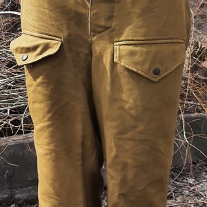 Op de afbeelding: Een bruine cargo broek met meerdere zakken en knoopsluitingen. De broek heeft een groene stoffen tailleband met een witte stoffen voering.