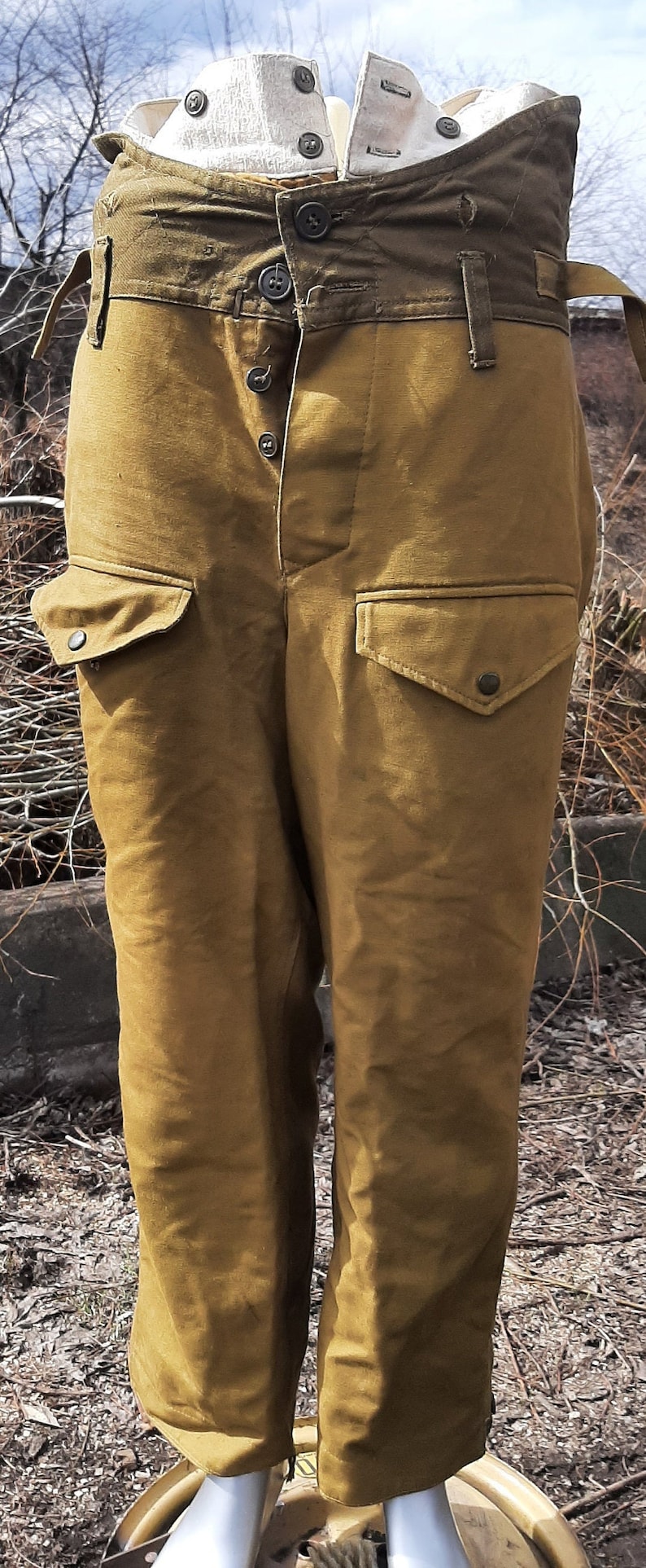 Op de afbeelding: Een bruine cargo broek met meerdere zakken en knoopsluitingen. De broek heeft een groene stoffen tailleband met een witte stoffen voering.