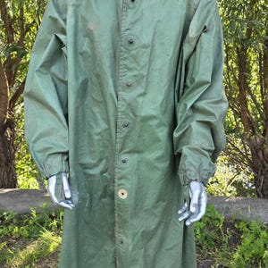 Op de afbeelding: Een lange, olijfgroene regenjas met een capuchon, knoopsluiting en lange mouwen. De jas wordt buiten op een mannequin getoond, met een natuurlijke groene achtergrond van bomen en gras. De jas lijkt gemaakt van een duurzaam, waterdicht materiaal.