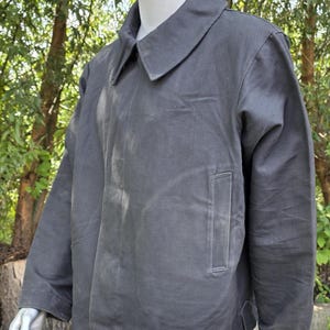 K&ouml;nnte beinhalten: Dunkelgraue Jacke mit Kragen und einer einzelnen Tasche auf der Vorderseite. Die Jacke wird auf einer Schaufensterpuppe pr&auml;sentiert. Der Hintergrund ist verschwommen, mit gr&uuml;nen B&auml;umen und Sonnenlicht.
