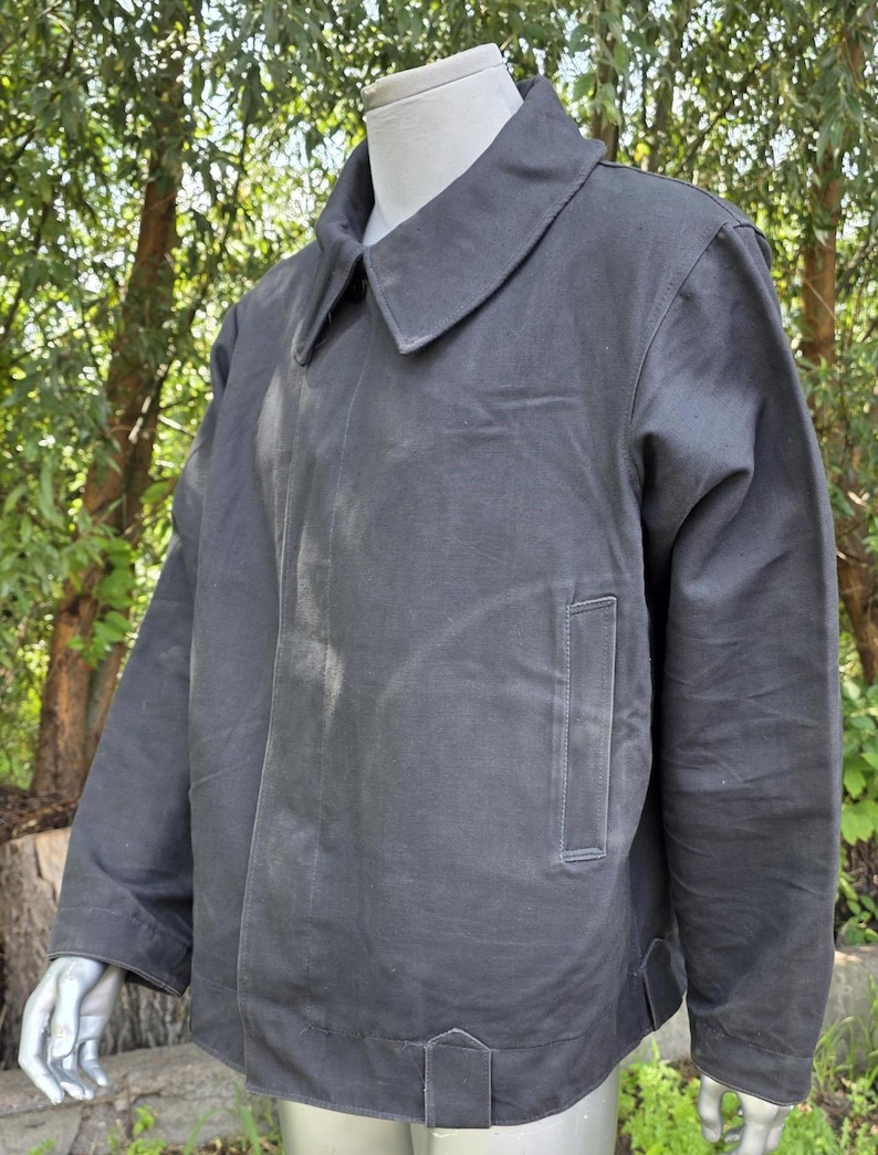 K&ouml;nnte beinhalten: Dunkelgraue Jacke mit Kragen und einer einzelnen Tasche auf der Vorderseite. Die Jacke wird auf einer Schaufensterpuppe pr&auml;sentiert. Der Hintergrund ist verschwommen, mit gr&uuml;nen B&auml;umen und Sonnenlicht.