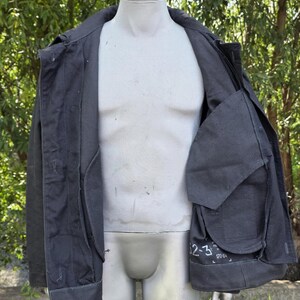 K&ouml;nnte beinhalten: Dunkelgraue Jacke mit Kragen und mehreren Taschen, pr&auml;sentiert auf einer Schaufensterpuppe. Die Jacke ist offen und zeigt das Innenfutter und eine gro&szlig;e Tasche mit wei&szlig;em Text. Die Jacke scheint aus strapazierf&auml;higem Stoff zu sein.