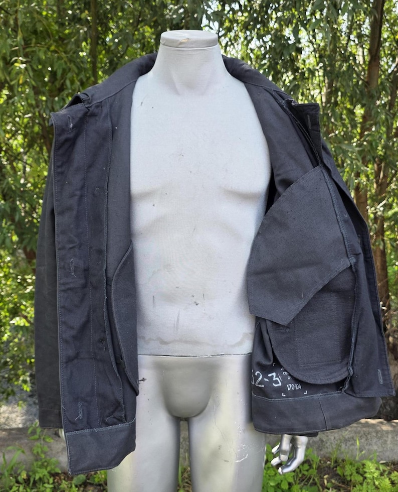 K&ouml;nnte beinhalten: Dunkelgraue Jacke mit Kragen und mehreren Taschen, pr&auml;sentiert auf einer Schaufensterpuppe. Die Jacke ist offen und zeigt das Innenfutter und eine gro&szlig;e Tasche mit wei&szlig;em Text. Die Jacke scheint aus strapazierf&auml;higem Stoff zu sein.