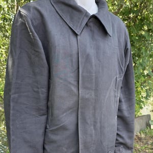 K&ouml;nnte beinhalten: Anthrazitgraue Jacke mit breitem Kragen und Knopfleiste. Die Jacke hat ein schlichtes, zweckm&auml;&szlig;iges Design mit geradem Saum und kleinen Laschen an den Manschetten. Der Hintergrund ist eine verschwommene Ansicht von gr&uuml;nem Laub.