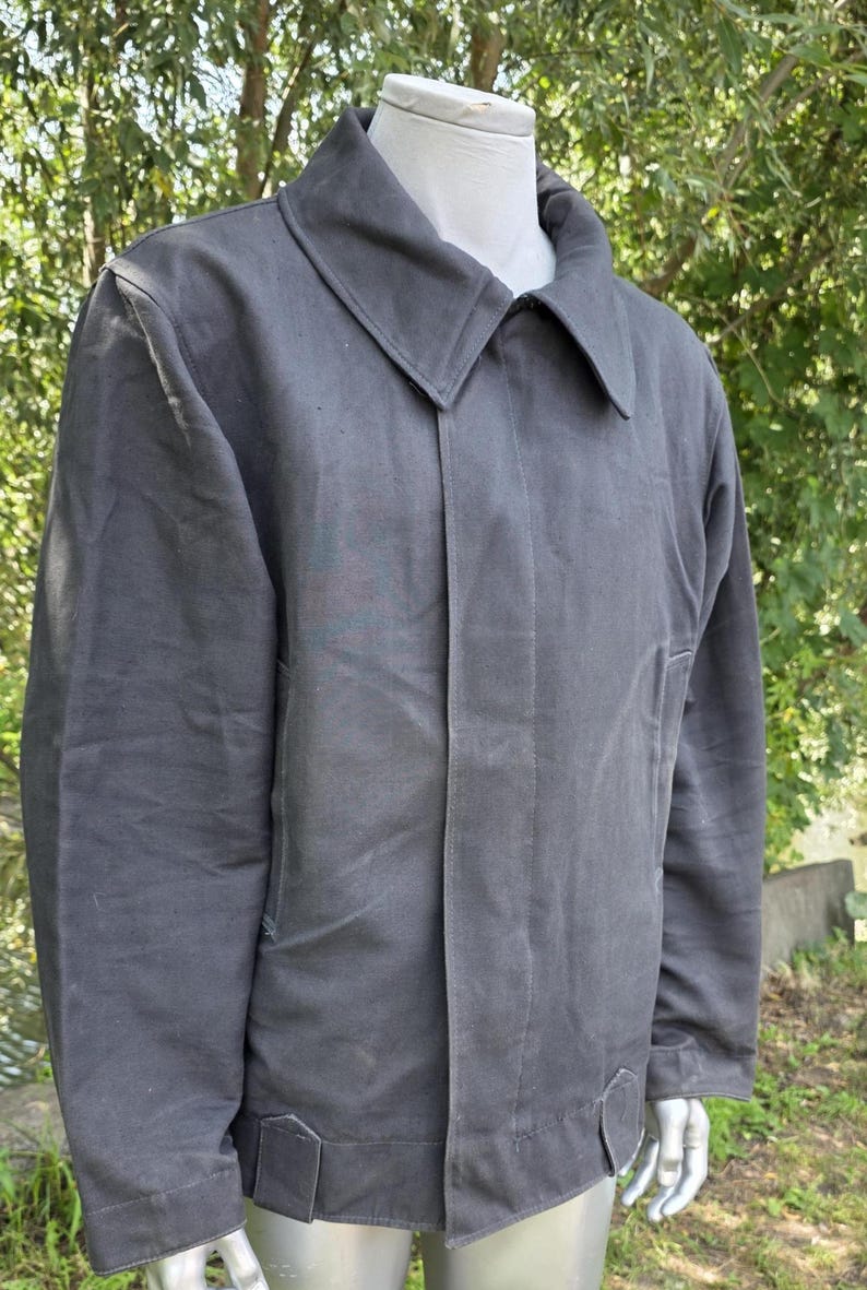K&ouml;nnte beinhalten: Anthrazitgraue Jacke mit breitem Kragen und Knopfleiste. Die Jacke hat ein schlichtes, zweckm&auml;&szlig;iges Design mit geradem Saum und kleinen Laschen an den Manschetten. Der Hintergrund ist eine verschwommene Ansicht von gr&uuml;nem Laub.