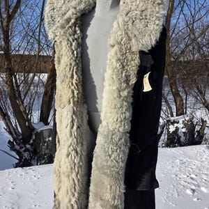 Puede incluir: Un abrigo largo negro con un grueso cuello y ribete de piel de oveja gris claro. El abrigo se exhibe en un maniquí en un entorno nevado al aire libre, con árboles desnudos y cielo azul de fondo.