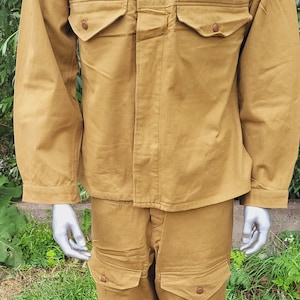 May include: A tan military uniform with a patch on the left breast featuring a yellow tank. The uniform includes a jacket and pants with multiple pockets.