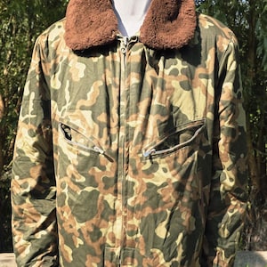 May include: Camouflage pattern jacket with a brown faux fur collar. The jacket has a zipper closure, two zippered pockets, and a button closure at the bottom. The jacket is a mix of green, brown, and tan colors.
