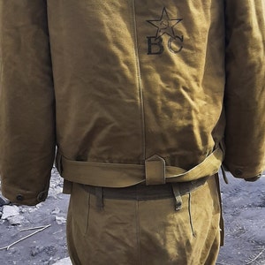 Op de afbeelding: Een bruin militair jack met een bont gevoerde capuchon en een ster en de letters "BC" geborduurd op de rug. Het jack heeft een riem en wordt gecombineerd met een bijpassende bruine broek.
