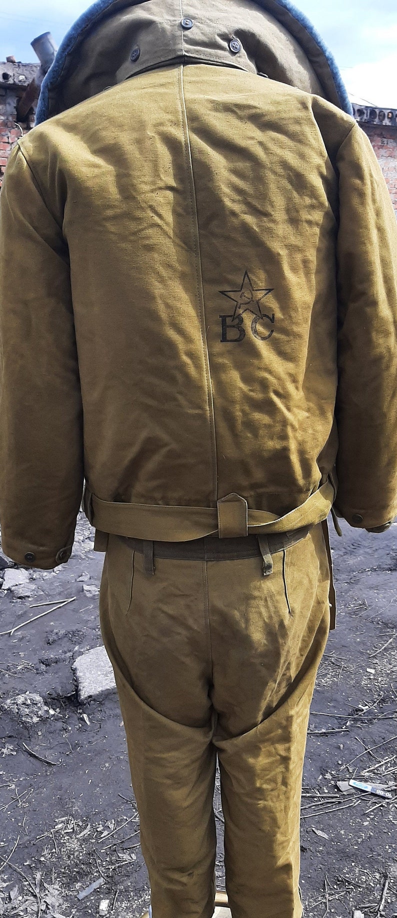 Op de afbeelding: Een bruin militair jack met een bont gevoerde capuchon en een ster en de letters "BC" geborduurd op de rug. Het jack heeft een riem en wordt gecombineerd met een bijpassende bruine broek.