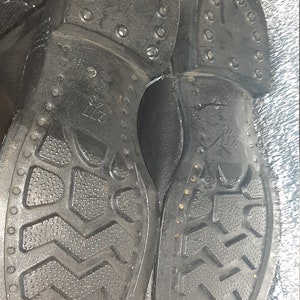 May include: Close-up of the soles of two black leather boots. The soles have a textured pattern and are studded with metal rivets. The boots are labeled "27 1/2".