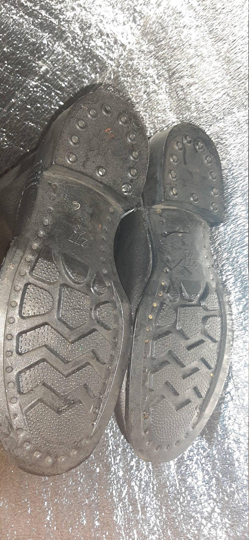 May include: Close-up of the soles of two black leather boots. The soles have a textured pattern and are studded with metal rivets. The boots are labeled "27 1/2".
