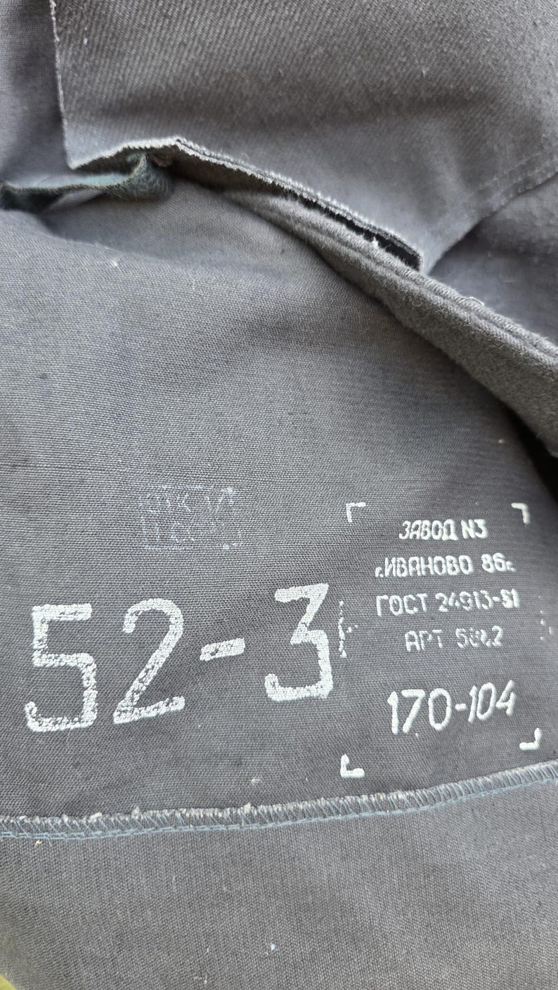 K&ouml;nnte beinhalten: Nahaufnahme eines grauen Stoffes mit wei&szlig;em Aufdruck und Zahlen. Die Zahlen "52-3" sind deutlich sichtbar. Zus&auml;tzlicher Text enth&auml;lt "ЗАВОД N3", "ИВАНОВО 86", "ГОСТ 24913-81", "APT 588.2" und "170-104".