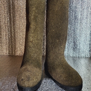 Puede incluir: Un par de botas de fieltro marrón con suelas de goma negra. Las botas son altas y tienen una punta redondeada.