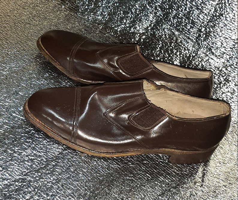 May include: A pair of brown leather slip-on shoes. The shoes have a cap toe design and elastic side panels. The soles show signs of wear. The shoes are likely vintage or pre-owned.