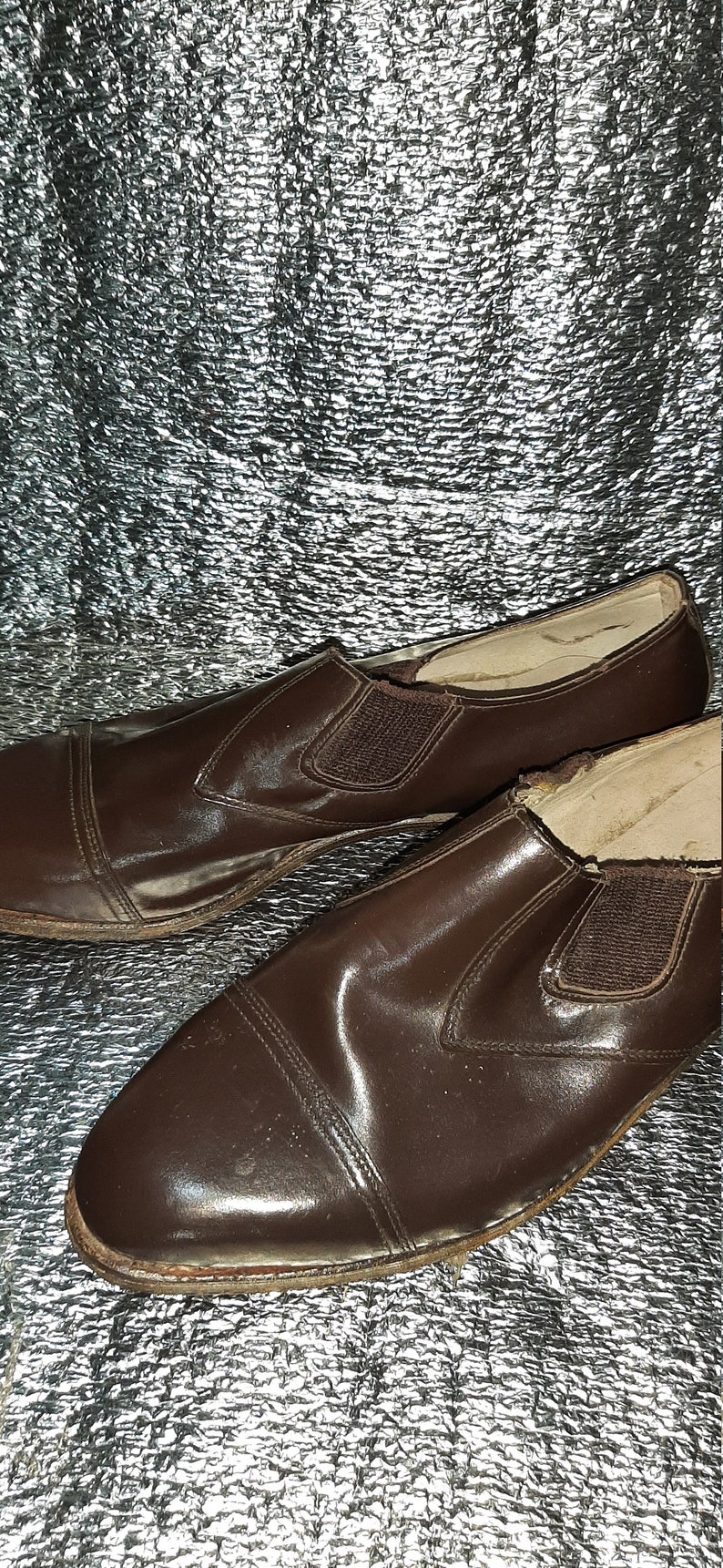 May include: A pair of brown leather slip-on shoes. The shoes have a cap toe design and elastic side panels. The leather has a glossy finish and shows signs of wear.