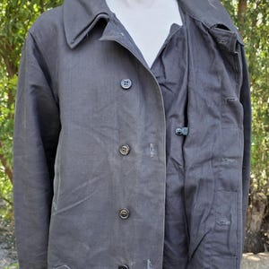 K&ouml;nnte beinhalten: Dunkelgraue Jacke mit Knopfleiste, Kragen und Taschen. Die Jacke wird auf einer Schaufensterpuppe pr&auml;sentiert, um ihr Design und ihre Passform zu zeigen. Der Stoff scheint ein strapazierf&auml;higes Material zu sein, das f&uuml;r verschiedene Wetterbedingungen geeignet ist.