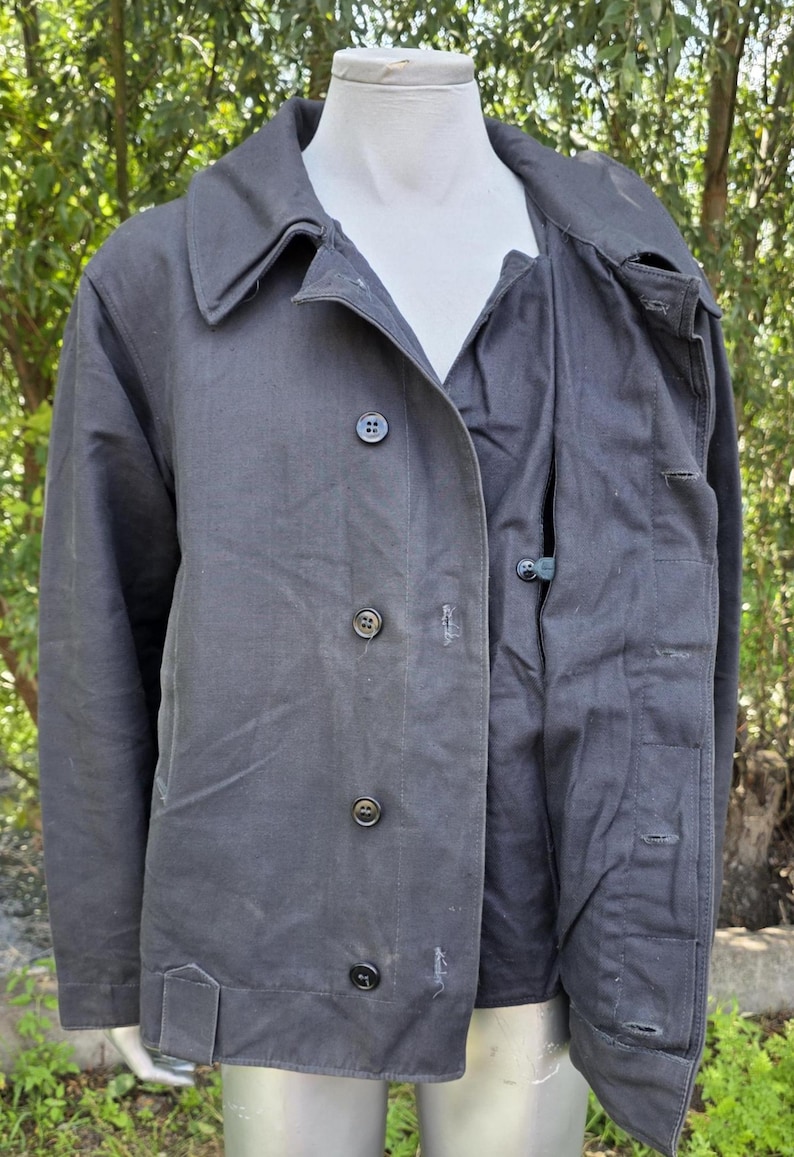 K&ouml;nnte beinhalten: Dunkelgraue Jacke mit Knopfleiste, Kragen und Taschen. Die Jacke wird auf einer Schaufensterpuppe pr&auml;sentiert, um ihr Design und ihre Passform zu zeigen. Der Stoff scheint ein strapazierf&auml;higes Material zu sein, das f&uuml;r verschiedene Wetterbedingungen geeignet ist.