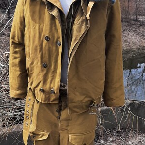 Op de afbeelding: Een bruin militair jack met een bontkraag en een bijpassende broek. Het jack heeft een dubbele rij knopen en een riem.