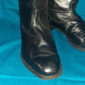 May include: A pair of black leather boots with a pointed toe and a low heel. The boots are on a blue background.