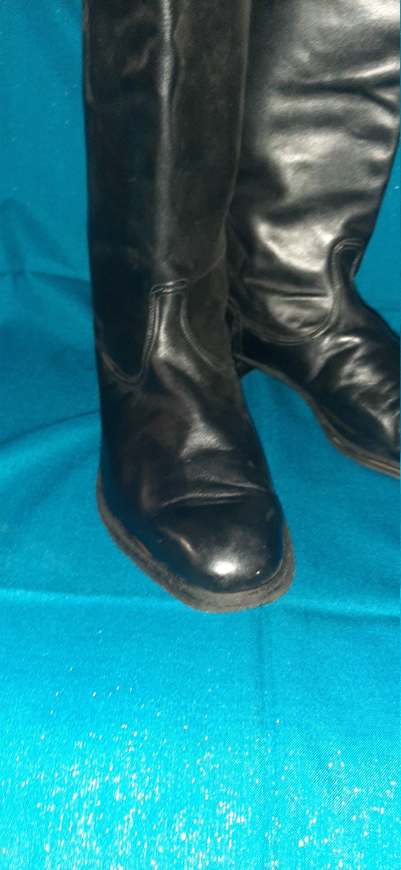 May include: A pair of black leather boots with a pointed toe and a low heel. The boots are on a blue background.