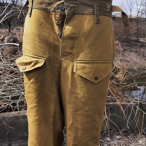 Op de afbeelding: Een paar bruine militaire broek met een groene canvas tailleband en meerdere knoopsluitingen. De broek heeft grote cargozakken aan de zijkanten.