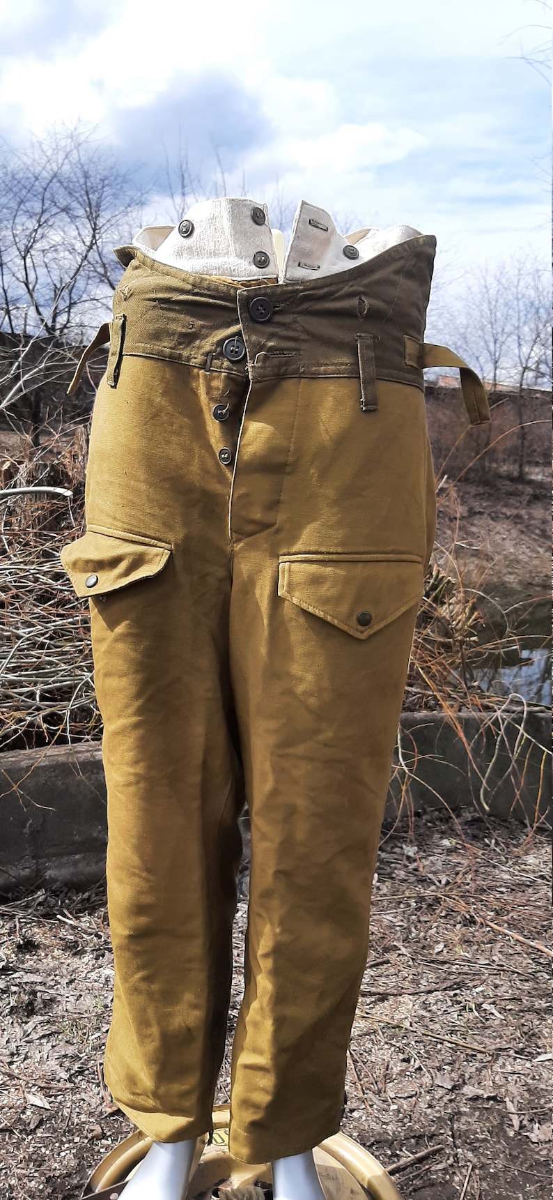 Op de afbeelding: Een paar bruine militaire broek met een groene canvas tailleband en meerdere knoopsluitingen. De broek heeft grote cargozakken aan de zijkanten.