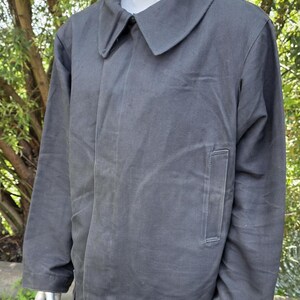 K&ouml;nnte beinhalten: Dunkelgraue Jacke mit breitem Kragen und einer einzelnen Tasche auf der Vorderseite. Die Jacke wird auf einer Schaufensterpuppe vor einem verschwommenen gr&uuml;nen Hintergrund pr&auml;sentiert. Die Jacke hat ein schlichtes, zweckm&auml;&szlig;iges Design.