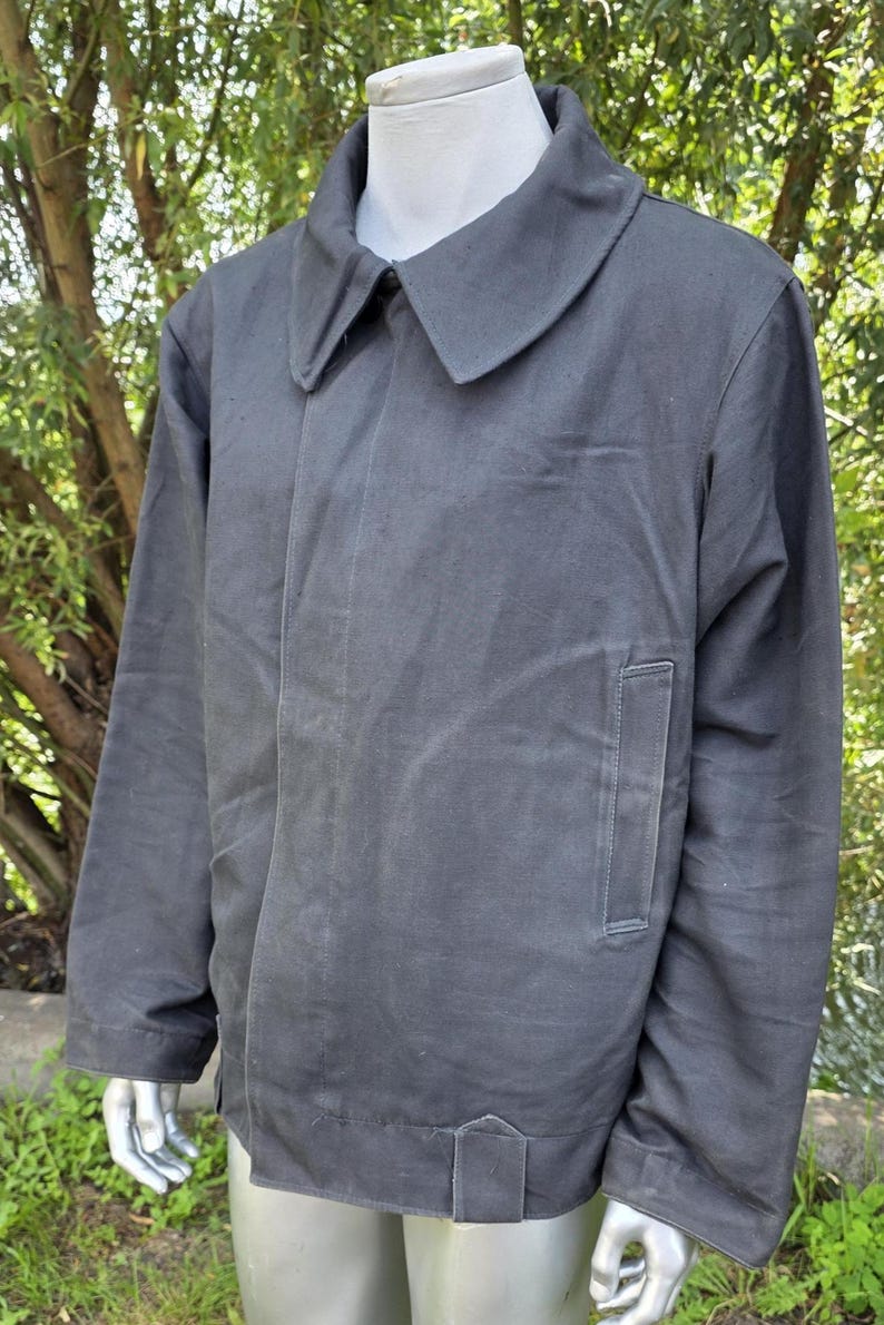 K&ouml;nnte beinhalten: Dunkelgraue Jacke mit breitem Kragen und einer einzelnen Tasche auf der Vorderseite. Die Jacke wird auf einer Schaufensterpuppe vor einem verschwommenen gr&uuml;nen Hintergrund pr&auml;sentiert. Die Jacke hat ein schlichtes, zweckm&auml;&szlig;iges Design.