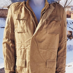 May include: A tan military-style jacket with multiple front pockets and a grey, fur-lined hood. The jacket is displayed on a mannequin in a snowy outdoor setting. The jacket has a button-up front.