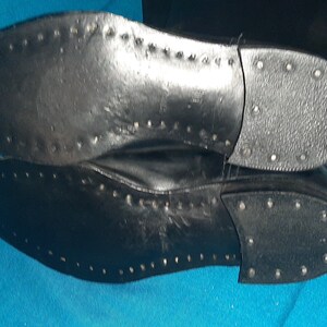 May include: Close-up of the black leather soles of a pair of boots. The soles have a textured pattern and are studded with small metal rivets.