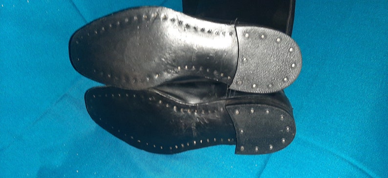 May include: Close-up of the black leather soles of a pair of boots. The soles have a textured pattern and are studded with small metal rivets.