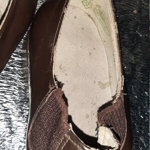May include: Close-up of a pair of brown leather shoes. The shoes have a slip-on design with elastic side panels. The interior of the shoe is visible, showing a worn insole with a logo.