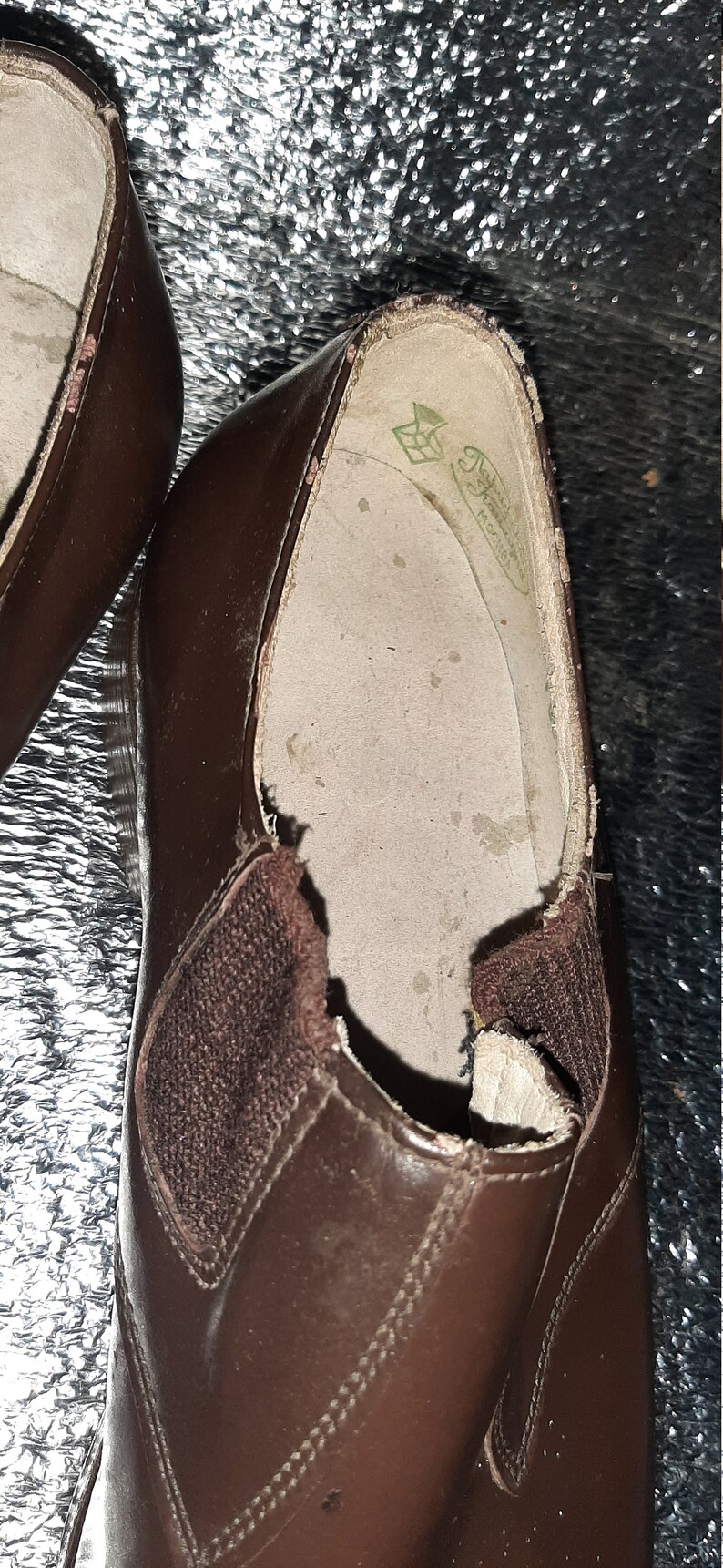 May include: Close-up of a pair of brown leather shoes. The shoes have a slip-on design with elastic side panels. The interior of the shoe is visible, showing a worn insole with a logo.