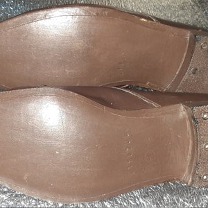 May include: A pair of brown leather shoes, soles facing up. The soles are a solid brown color, with a textured appearance. The heels are reinforced with metal studs.